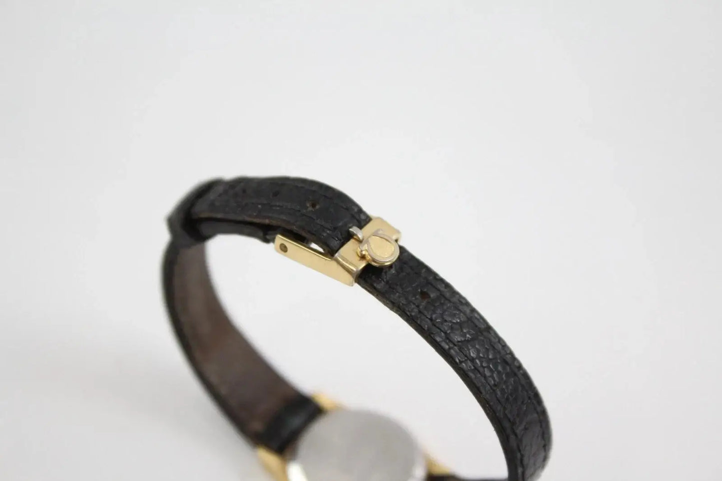 A close-up of a black textured leather watch band with gold-tone hardware and a silver-toned round case, shown against a plain white background.