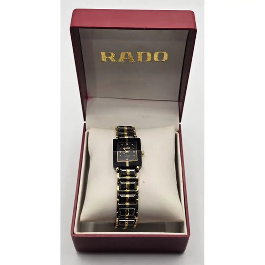 A RADO women's watch with a black rectangular dial and a black-and-gold link bracelet, presented inside its open dark red brand box with a cream cushion.