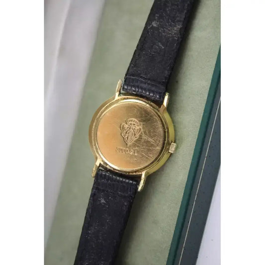 A vintage wristwatch with a round gold-tone case and a dark textured strap, shown resting on a light green cushion inside a box.