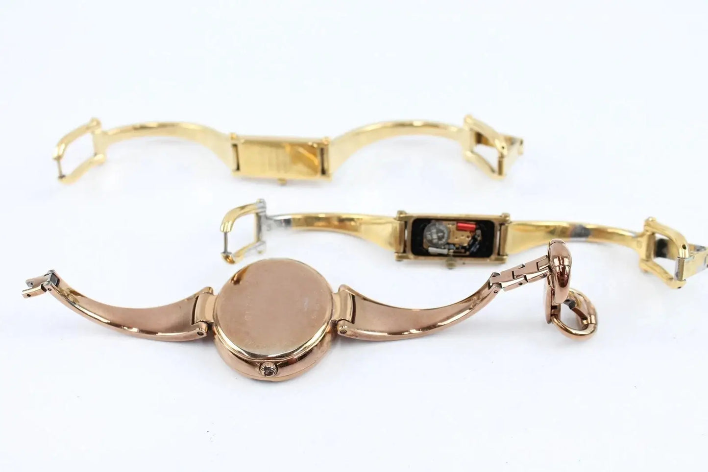 Two gold-tone wristwatches with bangle straps, shown from the back on a plain white background. One watch has a round case and the other has a rectangular case with a small colorful display window.