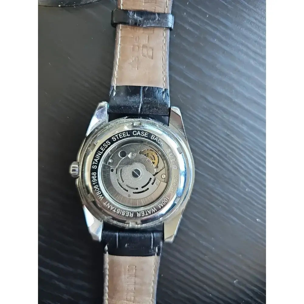 The back of a wristwatch with a silver stainless steel case and a light brown leather strap, showing the automatic mechanical movement through a transparent case cover.