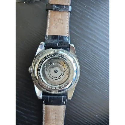 The back of a wristwatch with a silver stainless steel case and a light brown leather strap, showing the automatic mechanical movement through a transparent case cover.