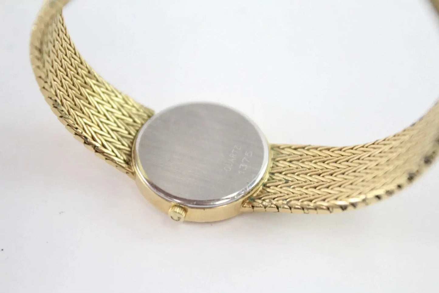 Close-up of a gold-tone mesh bracelet watch showing the flat round back cover and side crown, on a plain white background.