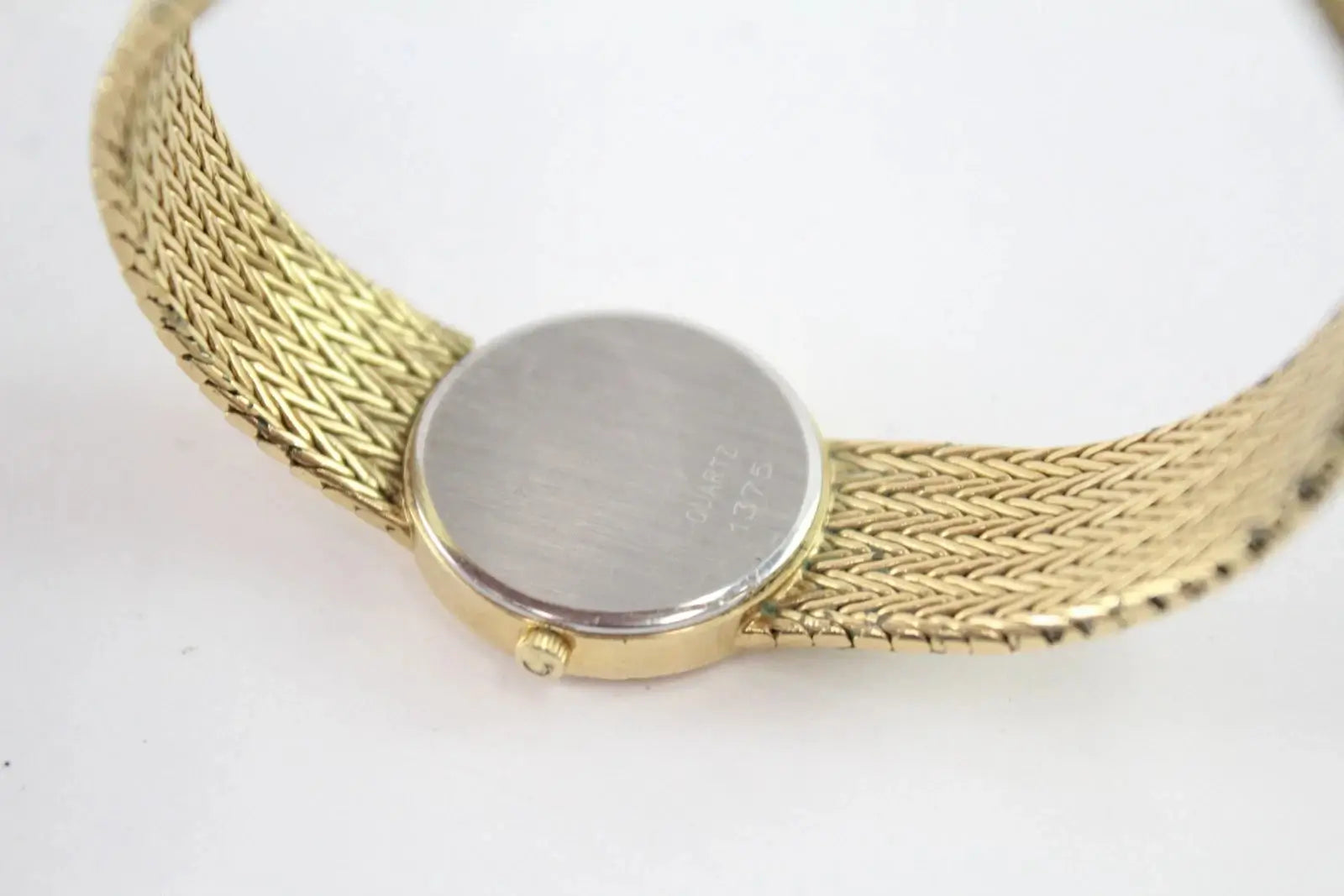 Close-up of a gold-tone mesh bracelet watch showing the flat round back cover and side crown, on a plain white background.