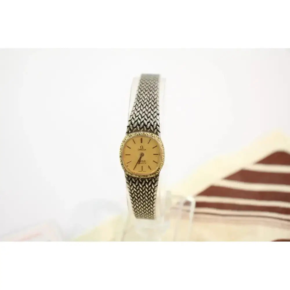 A vintage Omega women's watch with a gold dial and a silver-and-black chevron-patterned metal bracelet, displayed on a clear stand against a white background.