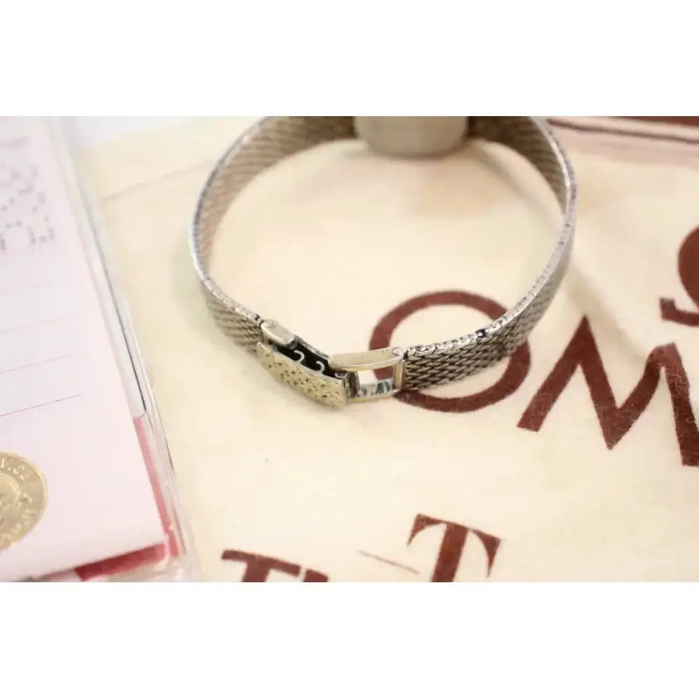 A silver-tone metal mesh bracelet with a sliding clasp, displayed on a cream-colored branded fabric surface.
