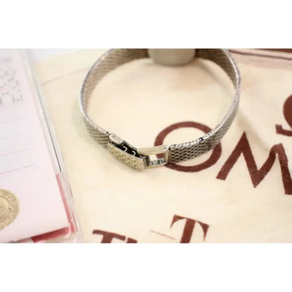 A silver-tone metal mesh bracelet with a sliding clasp, displayed on a cream-colored branded fabric surface.