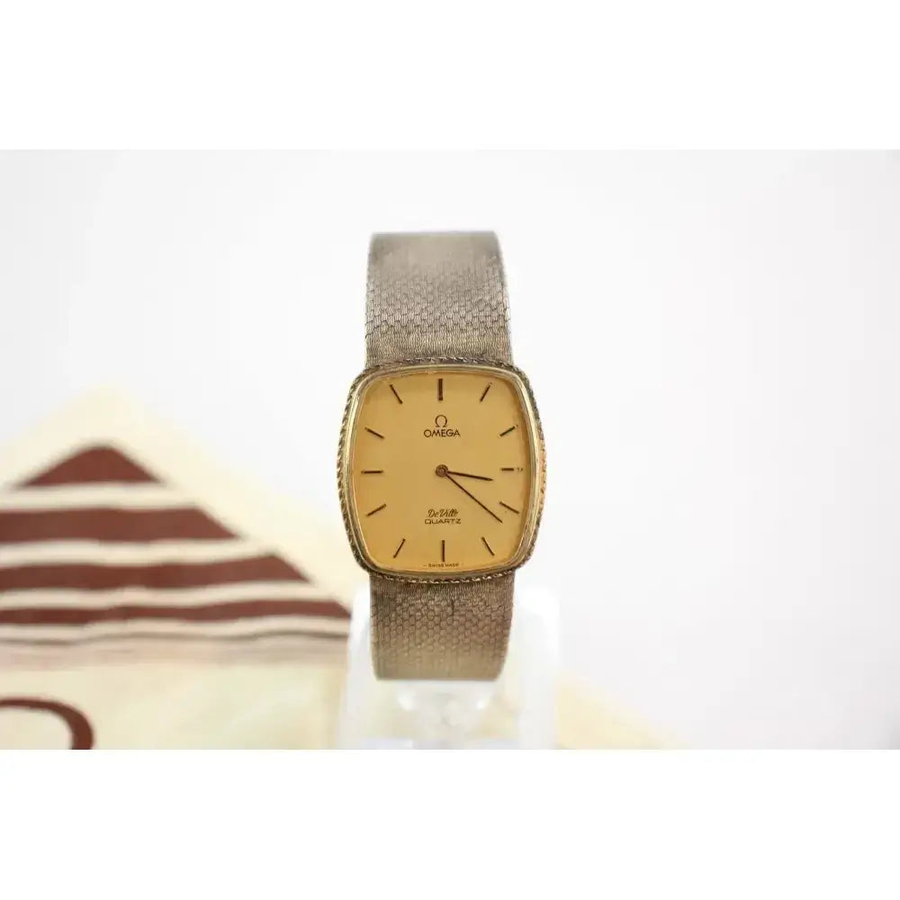 An Omega S-Prism watch with a gold-toned dial and a textured beige mesh strap, displayed on a clear stand against a white background.