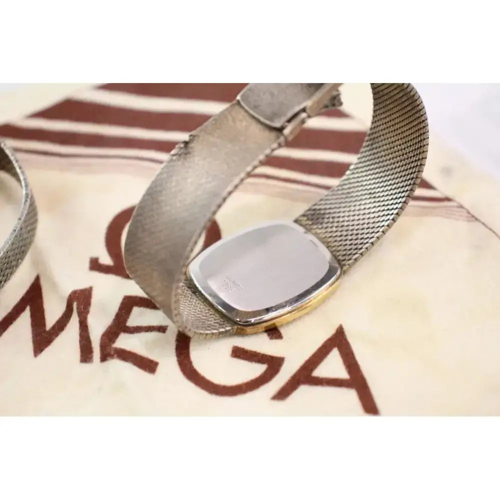 A close-up of a vintage Omega watch with a silver-tone mesh band and a gold-tone bezel, resting on its cream-colored Omega packaging box.