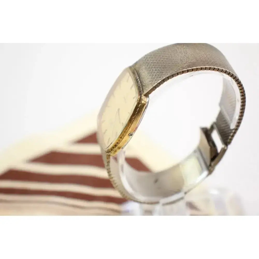 A vintage-style wristwatch with a textured silver-tone metal mesh band and a gold-tone case, shown on a clear display stand against a white background.