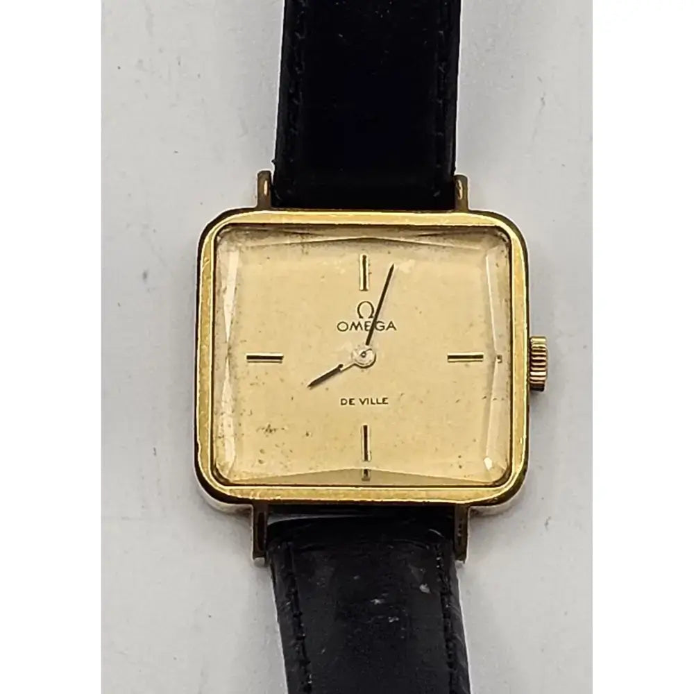 Omega De Ville vintage wristwatch with a gold-tone square case, cream-colored dial, and black leather strap, shown against a light gray background.