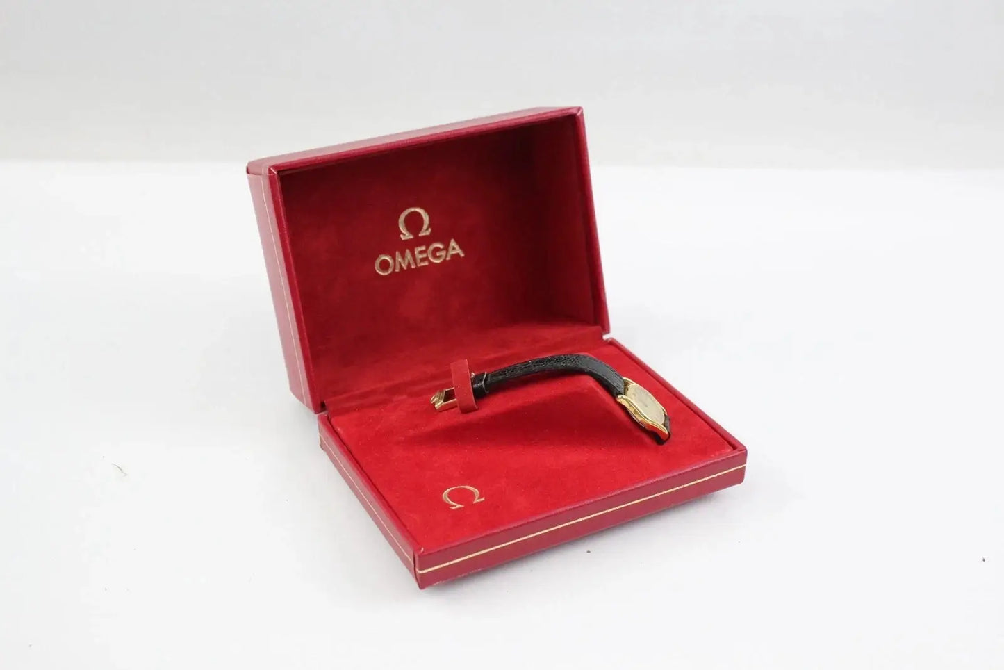 An Omega wristwatch with a black strap and gold-toned case, presented inside its open red Omega box on a white background.