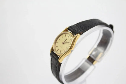 A vintage-style wristwatch with a gold-tone oval case, cream dial, and textured black leather strap, displayed on a clear stand against a white background.