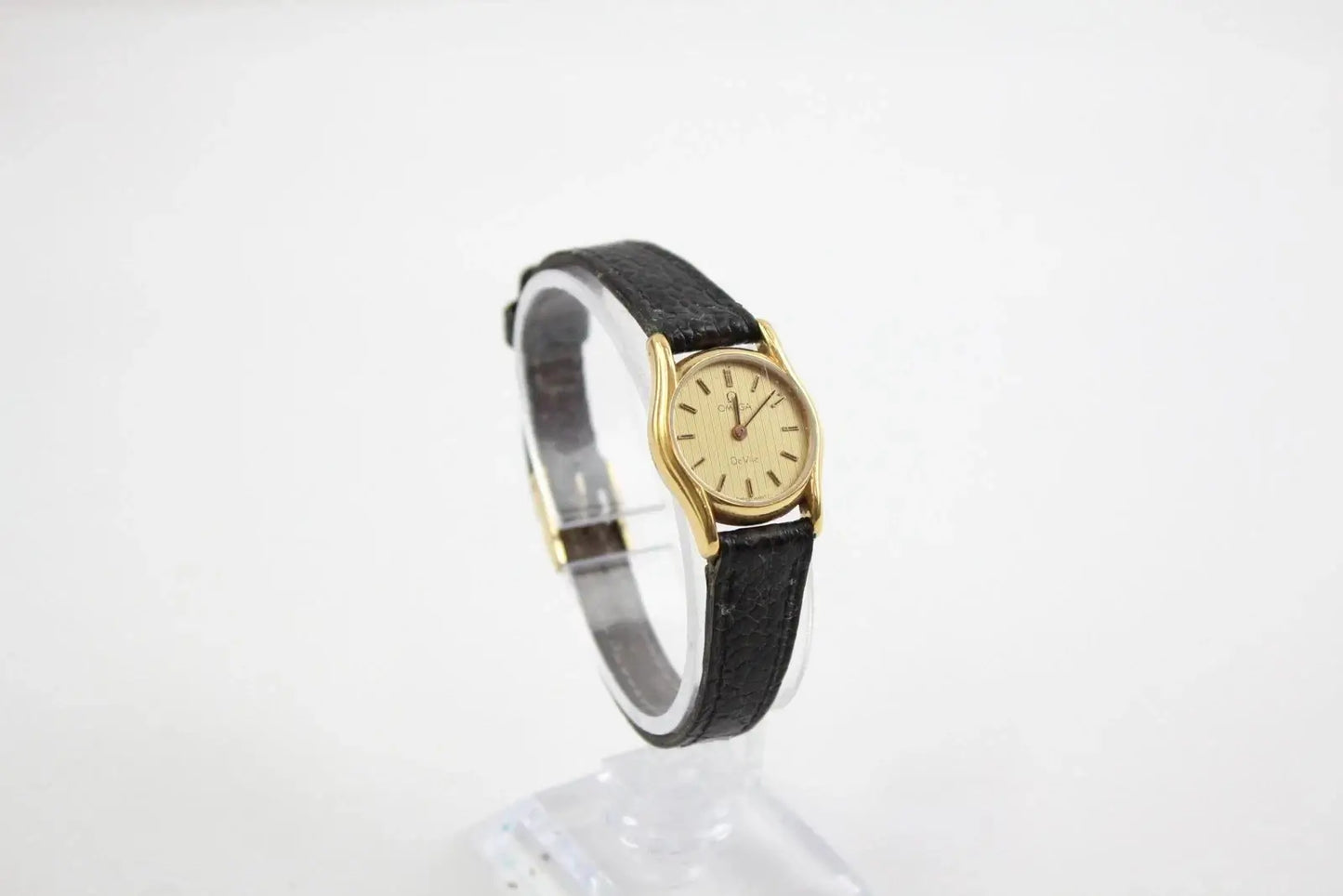 A vintage-style wristwatch with a gold-tone oval case, cream dial, and black leather strap, displayed on a clear stand against a white background.