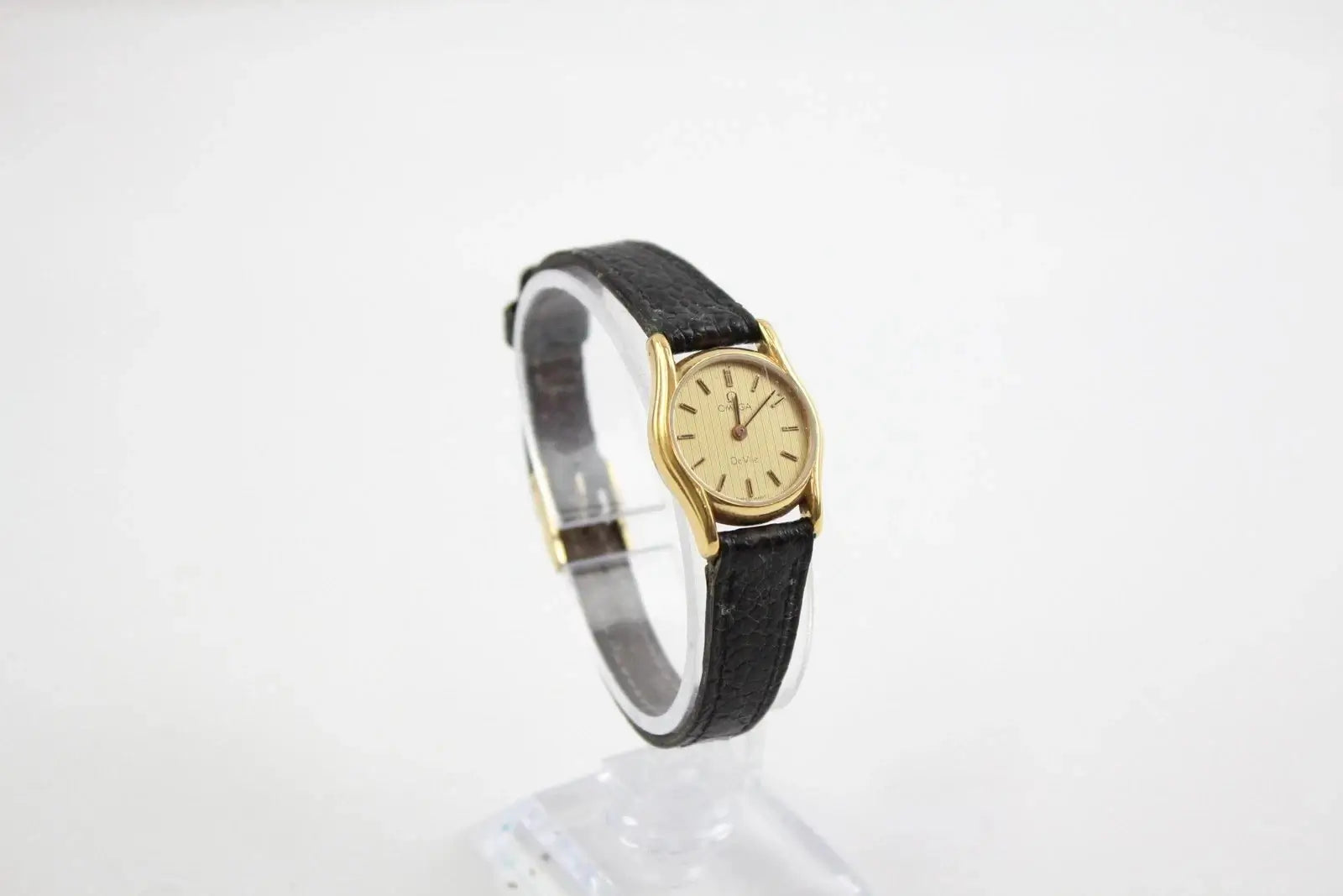 A vintage-style wristwatch with a gold-tone oval case, cream dial, and black leather strap, displayed on a clear stand against a white background.