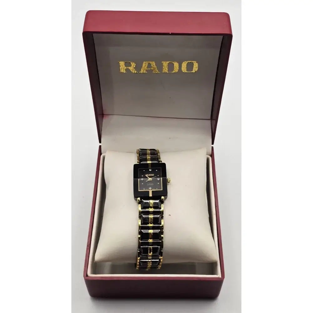 A RADO women's watch with a black rectangular dial and a black-and-gold link bracelet, presented inside its open dark red brand box with a cream cushion.