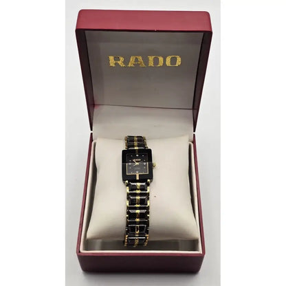 A RADO women's watch with a black rectangular dial and a black-and-gold link bracelet, presented inside its open dark red brand box with a cream cushion.