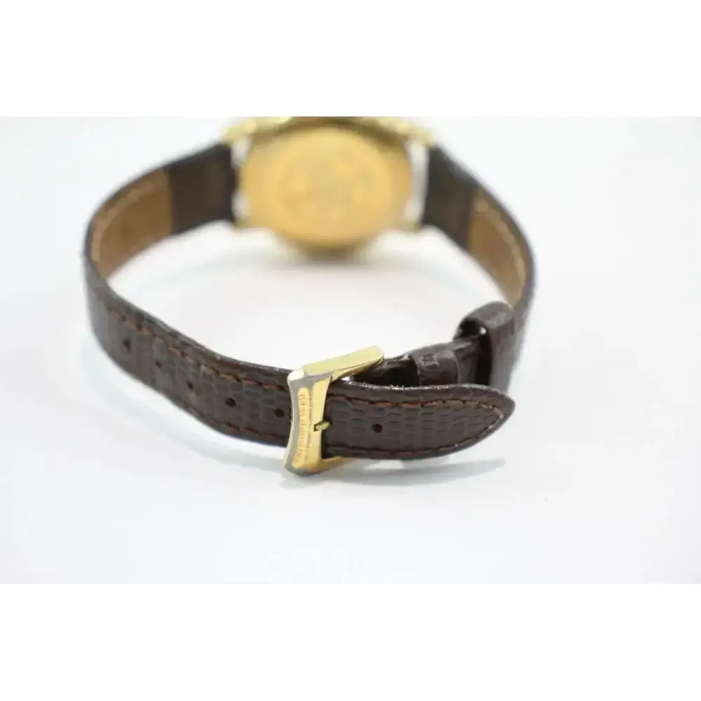 A close-up of a watch strap with a textured dark brown leather finish and a gold-tone buckle, shown from the back against a plain white background.