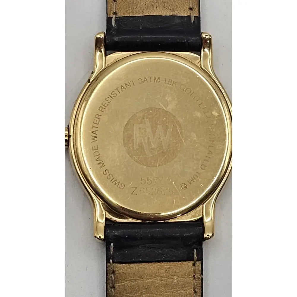 The back of a wristwatch with a gold-tone case and a black leather strap, showing engraved text and a monogram logo on the gold-toned backplate.