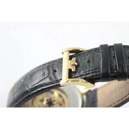 Close-up of a black alligator leather watch strap with a gold H-shaped buckle, showing the textured leather and a glimpse of the gold-toned watch case.