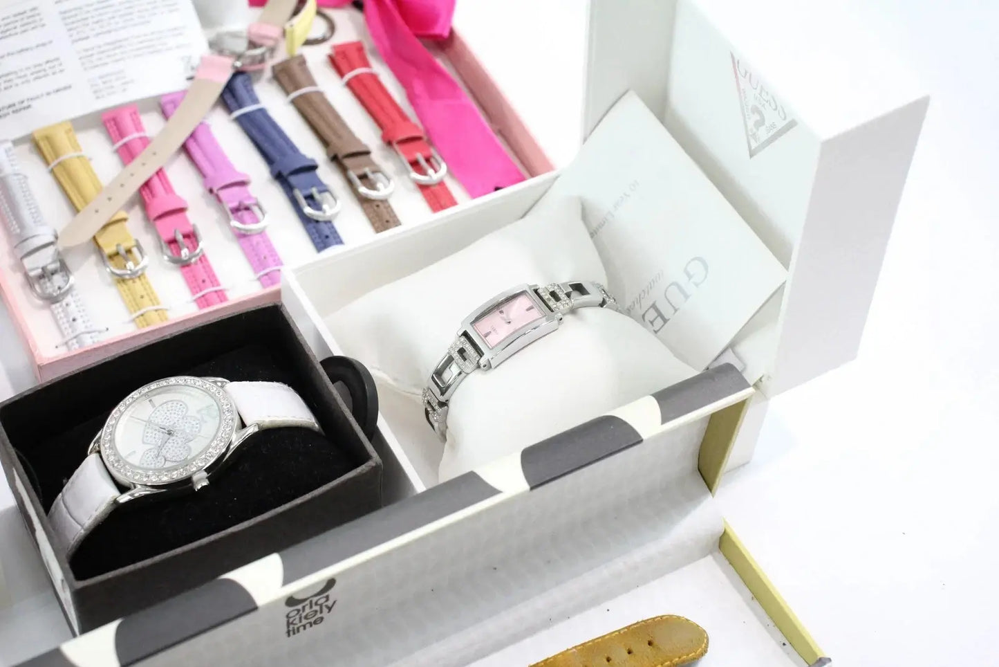 A Gucci watch with a silver-toned metal bracelet and a light pink dial, displayed inside an open white Gucci box. Another view of the same watch with a white strap is shown in a black box.