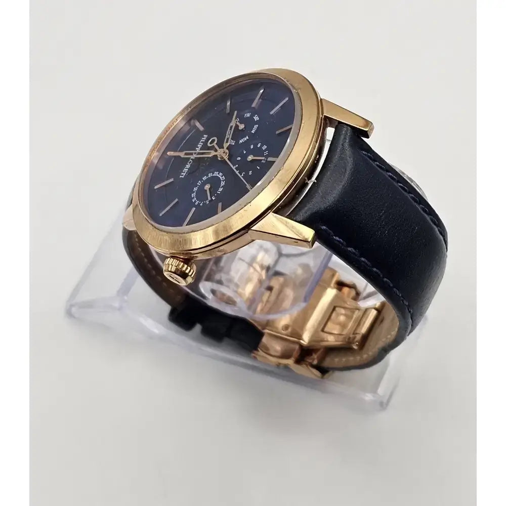 A luxury wristwatch with a round rose gold case, a dark blue dial featuring moonphase indicators, and a black leather strap, displayed on a clear acrylic stand against a light gray background.