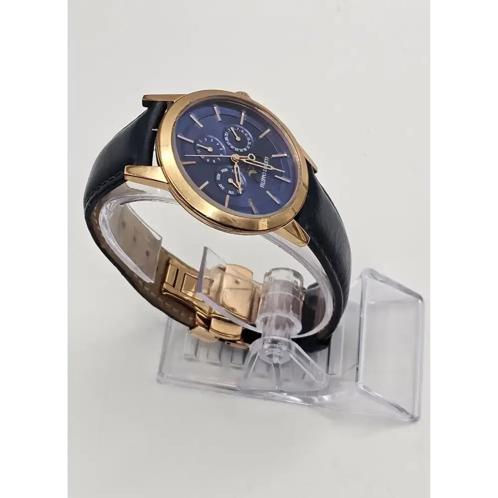 A men's luxury watch with a blue dial, gold-tone case, and black leather strap, displayed on a clear plastic stand against a light gray background.