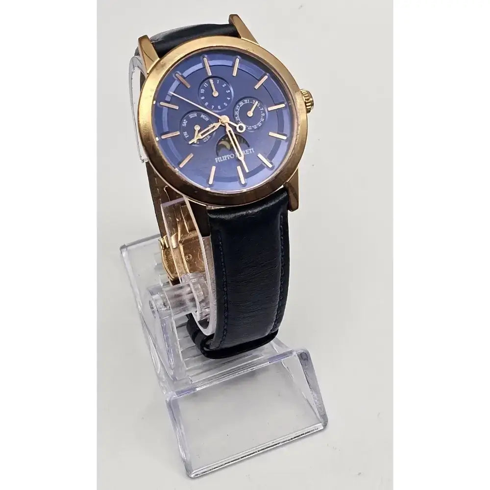 A men's analog watch with a round blue dial, gold-tone case, and black leather strap, displayed on a clear acrylic stand against a light gray background.