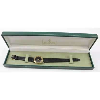 A Gucci wristwatch with a black leather strap and gold-tone case, presented inside its original green and beige Gucci box.