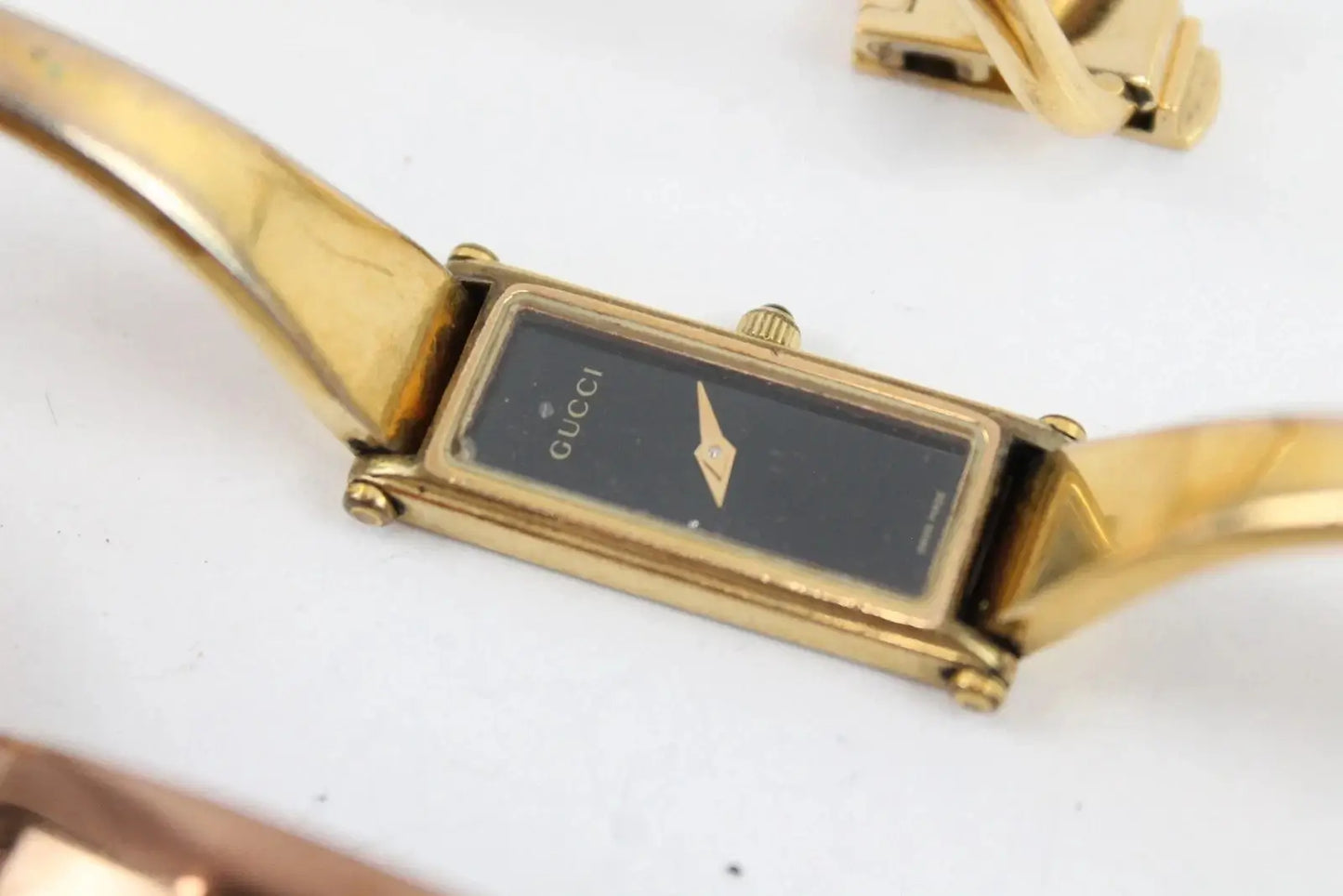 A close-up of a Gucci gold-tone watch with a black rectangular dial and gold-tone metal bracelet on a white background.
