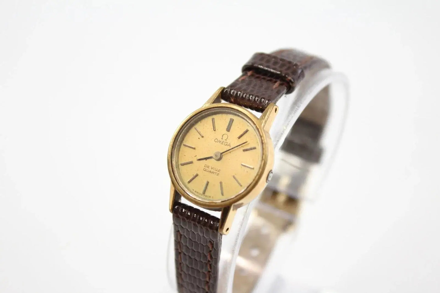 Omega wrist watch with a gold-tone case, gold dial, and brown textured leather strap, displayed on a clear stand against a white background.