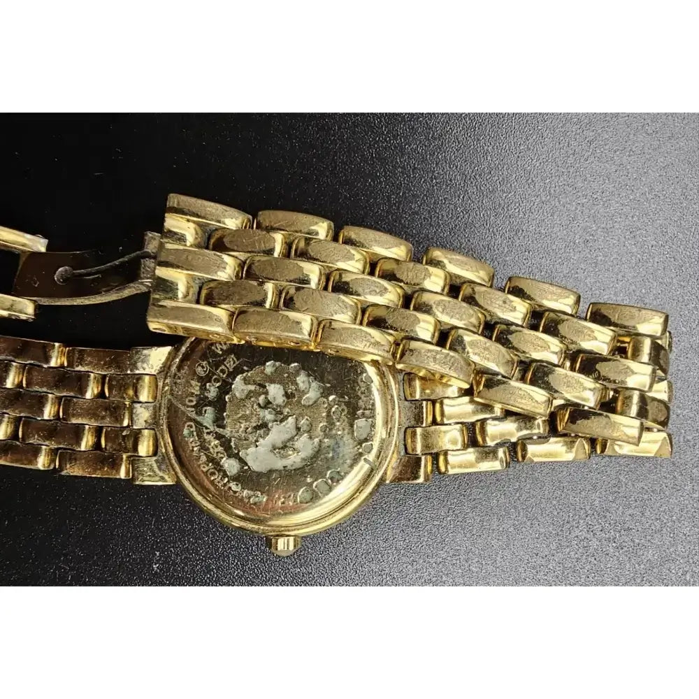 A close-up of a gold-tone metal watch band with a round case, photographed on a dark textured background. The watch face is visible through the open back, and the clasp is shown on the left.