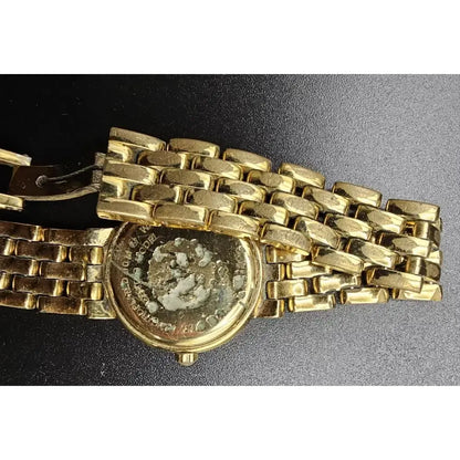 A close-up of a gold-tone metal watch band with a round case, photographed on a dark textured background. The watch face is visible through the open back, and the clasp is shown on the left.