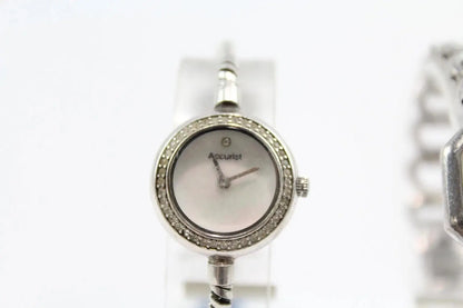 A silver-tone Accuulet women's watch with a round bezel set with small clear stones and a silver dial, shown on a white display stand.