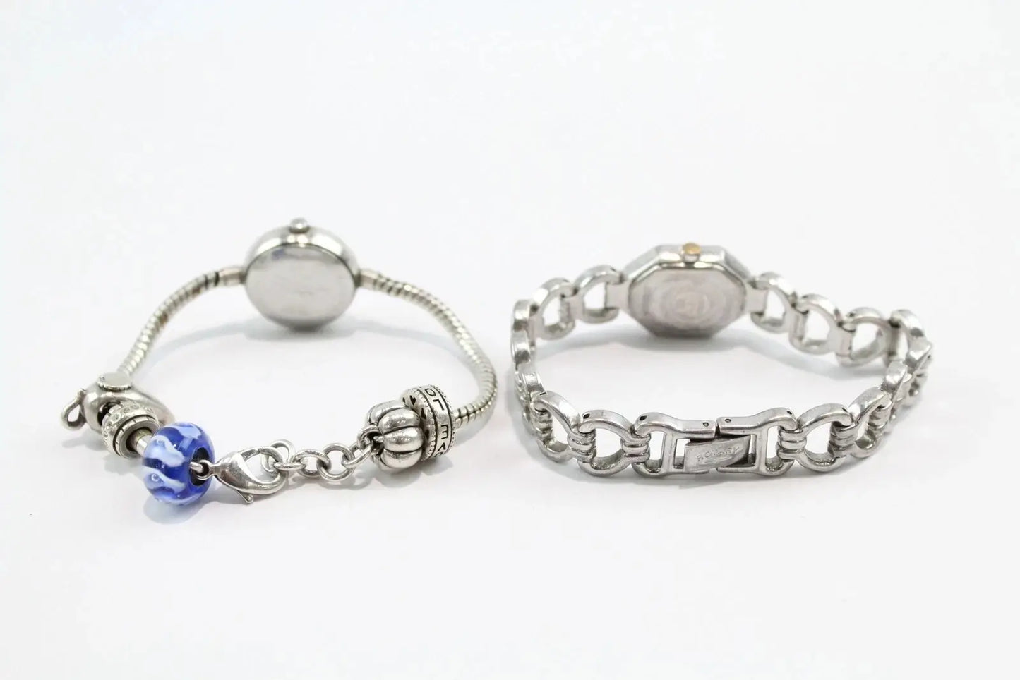 Two silver-toned bracelets displayed side by side on a white background. The left bracelet is a delicate snake chain with a round silver bead, a decorative filigree charm, and a blue faceted bead.