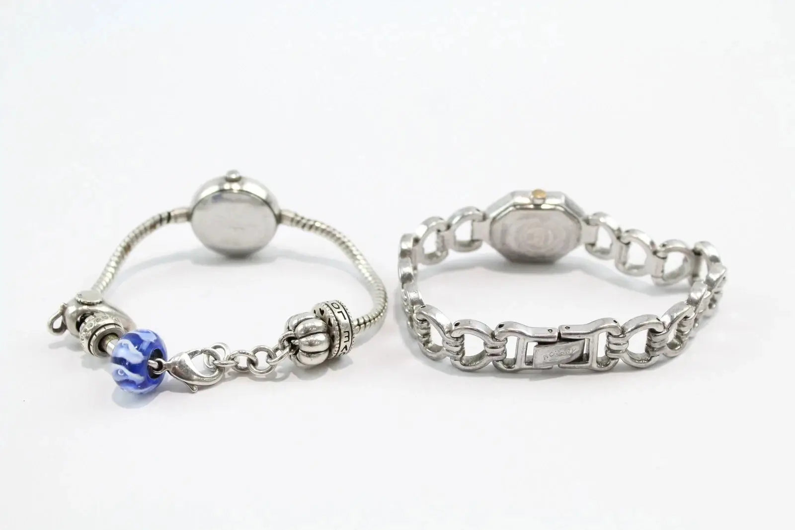 Two silver-toned bracelets displayed side by side on a white background. The left bracelet is a delicate snake chain with a round silver bead, a decorative filigree charm, and a blue faceted bead.