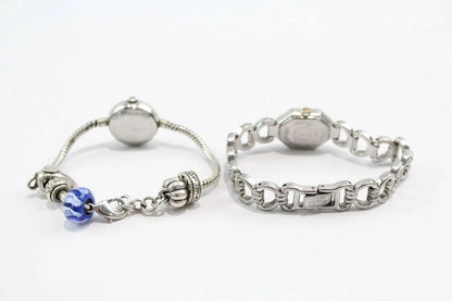 Two silver-toned bracelets displayed side by side on a white background. The left bracelet is a delicate snake chain with a round silver bead, a decorative filigree charm, and a blue faceted bead.